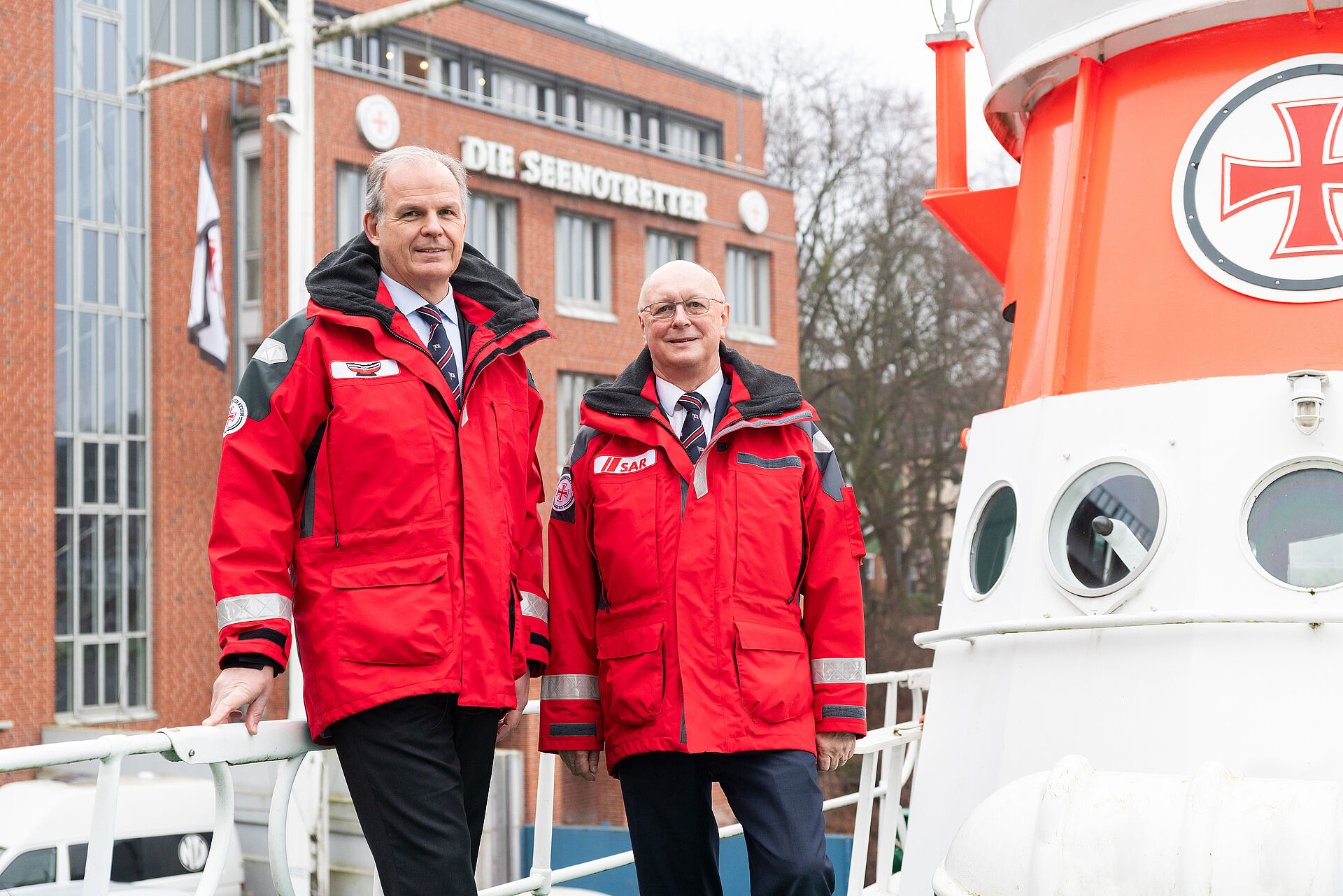 Die beiden Geschäftsführer stehen in roten Wetterjacken vor dem Geländer eines historischen Seenotrettungskreuzer. Im Hintergrund steht das Gebäude der Zentrale der Seenotretter.