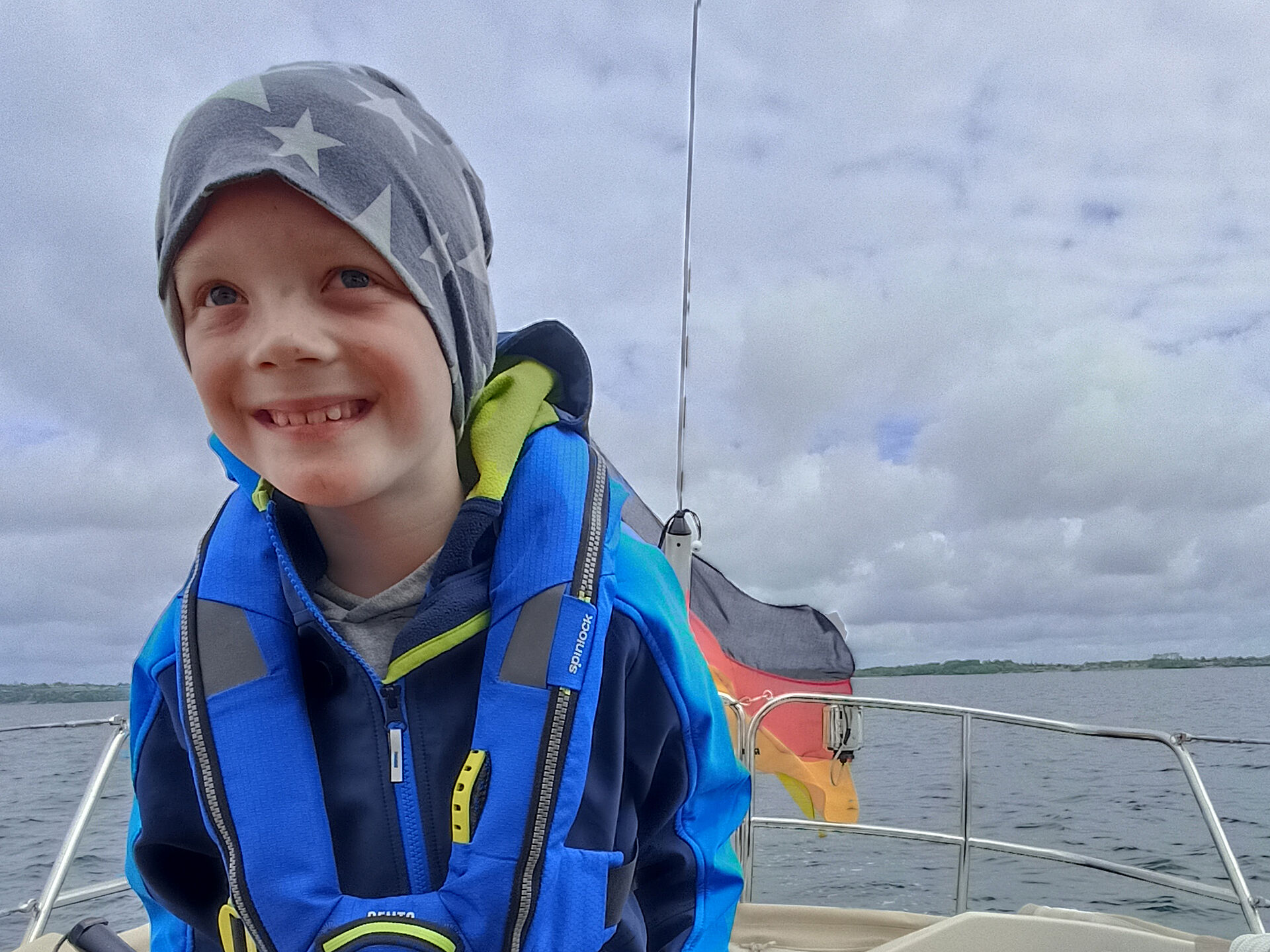 Neunjähriger Junge mit blauer Rettungsweste und grauem Stirnband bekleidet an Bord eines Segelbootes vor Wasser und bedecktem Himmel. 