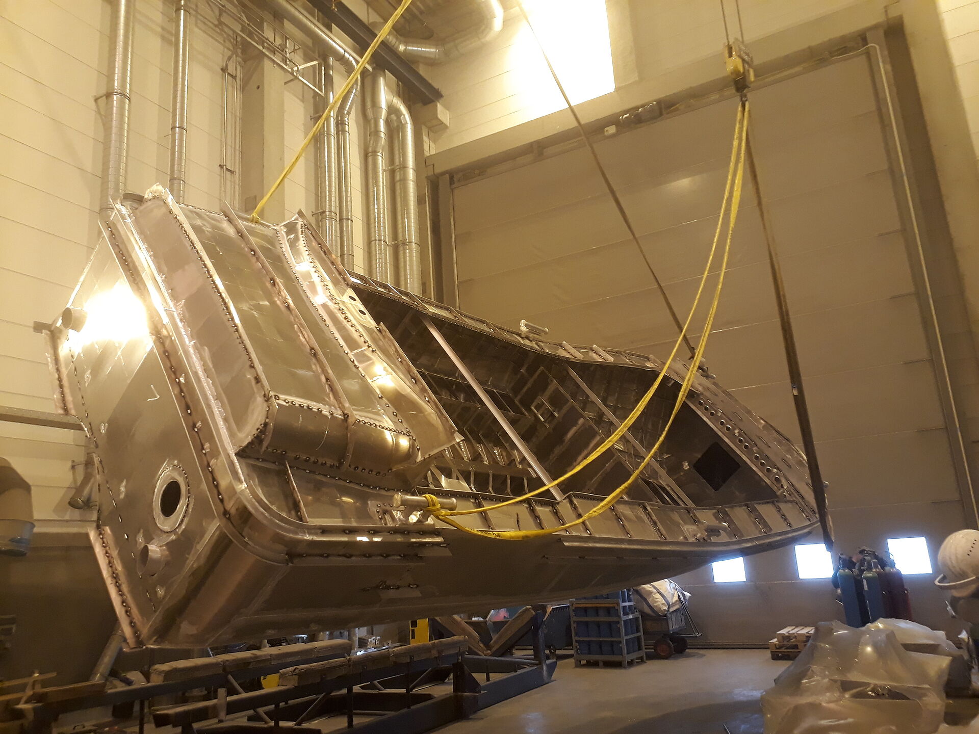 Der Rohbau aus Aluminium eines Seenotrettungsbootes wird in einer Werfthalle von einem Hallenkran um die Längsachse gedreht. Das Boot ist fast wieder aufgerichtet.