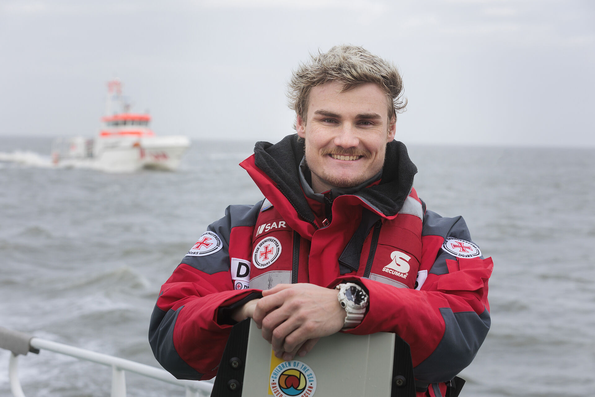 Kiteprofi Linus Erdmann steht mit seinem Board an der Reling eines Seenotrettungskreuzers, im Hintergrund ist der Seenotrettungskreuzer ANNELIESE KRAMER in Fahrt zu sehen.