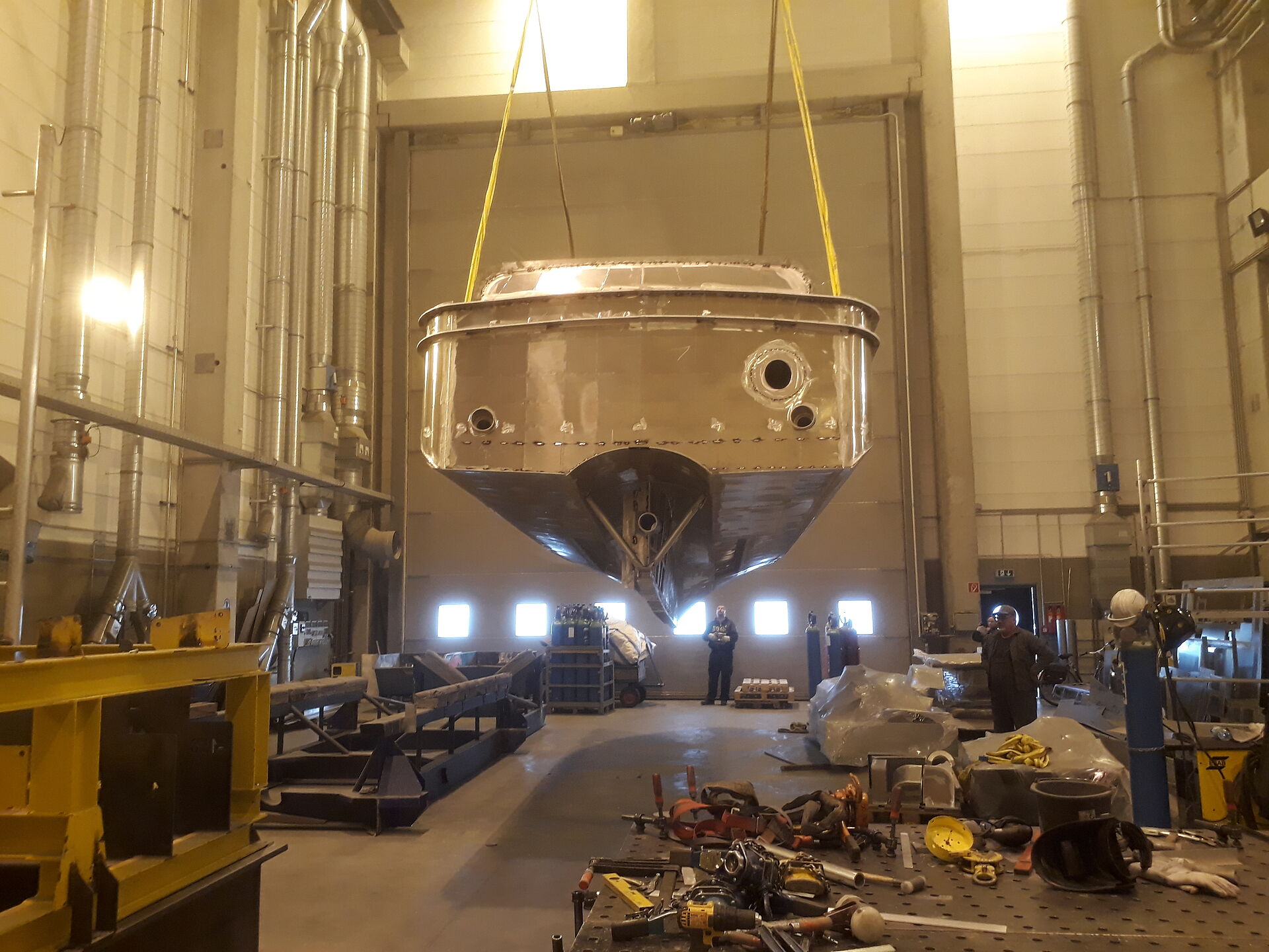 Der Rohbau aus Aluminium eines Seenotrettungsbootes wird in einer Werfthalle von einem Hallenkran gehoben. Ansicht von hinten, während das Boot einige Meter über dem Hallenboden schwebt.