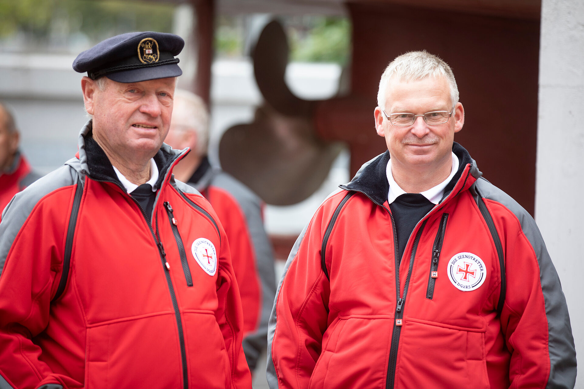 Zwei Seenotretter stehen entspannt in roter Einsatzkleidung vor der Schraube eines historischen Seenotrettungskreuzers.