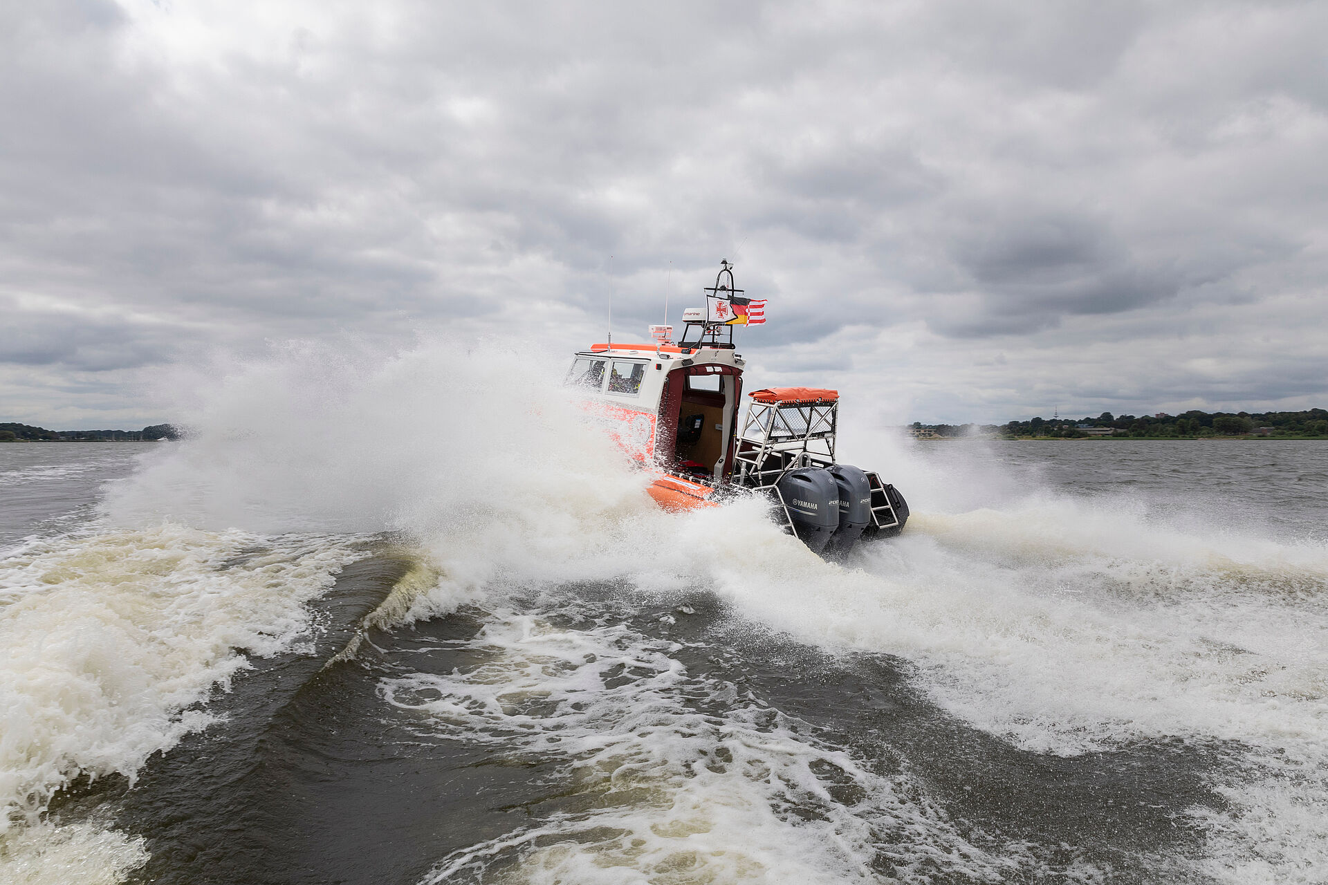Heckansicht des Seenotrettungsbootes HERWIL GÖTSCH: Es fährt in die aufgewühlten Wellen, Wasser spritzt seitlich vom Rumpf des Bootes weg.