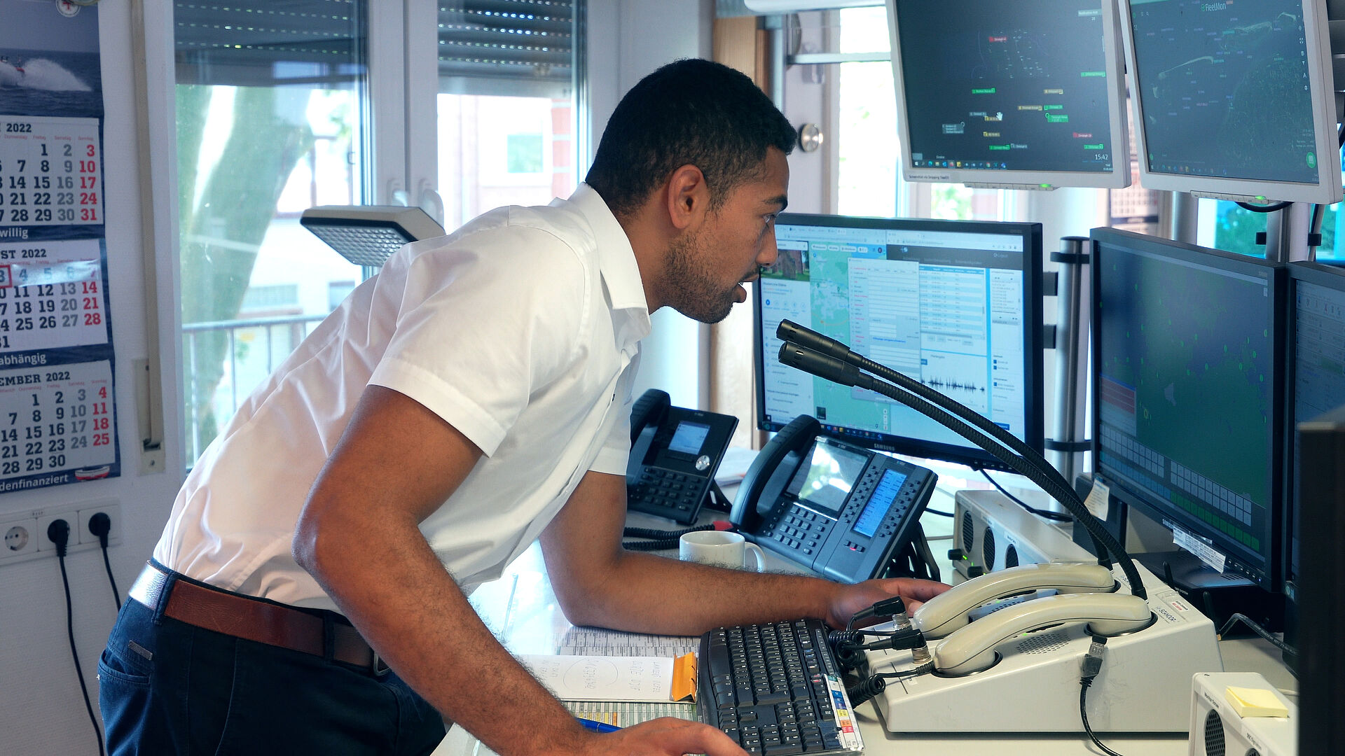 Ein junger Mann beugt sich zu einem Funkhörer vor mehreren Bildschirmen, die elektronische Seekarten zeigen.