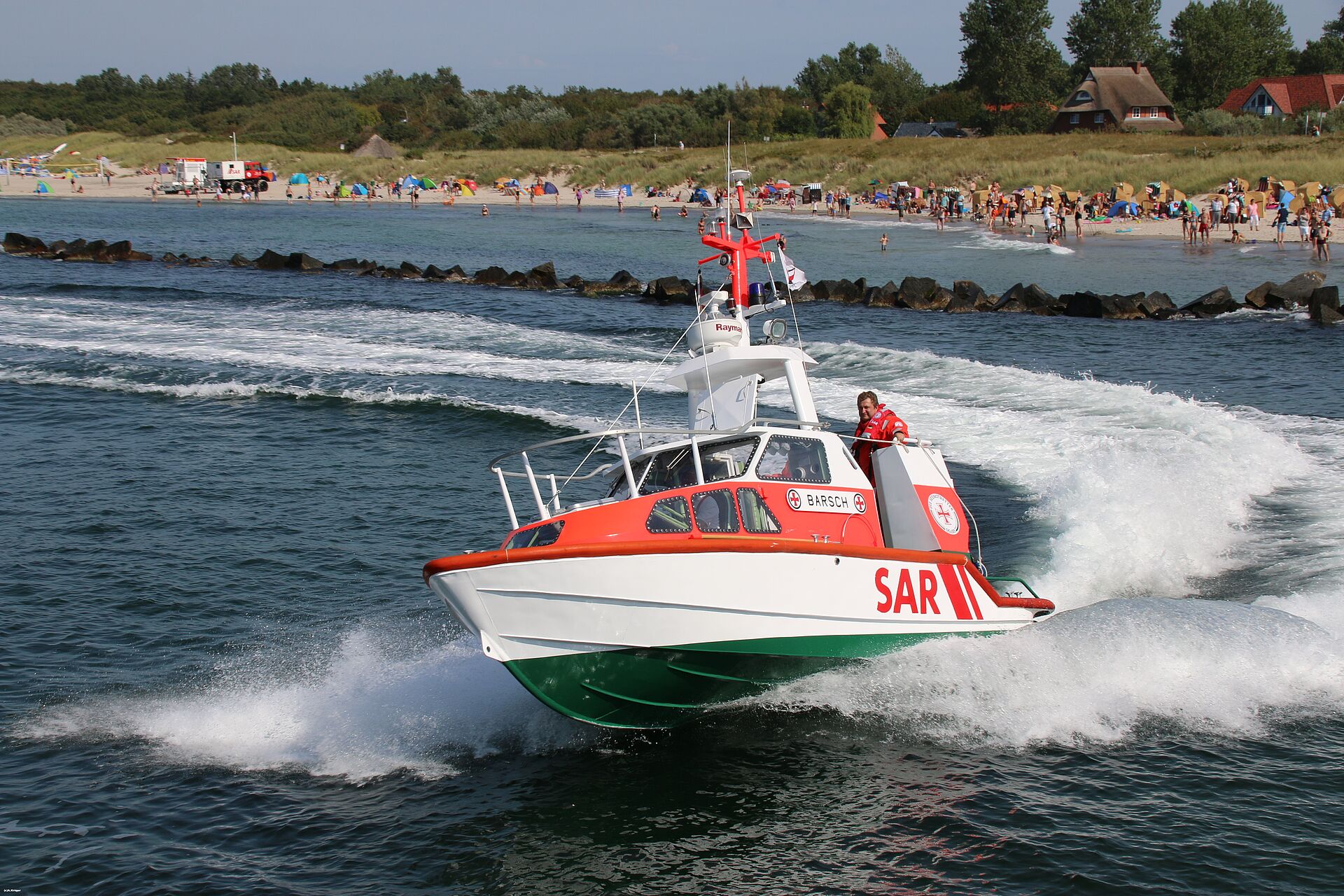 Das Seenotrettungsboot BARSCH fährt in schneller Fahrt auf der Ostsee vor einem Strand eine Kurve.