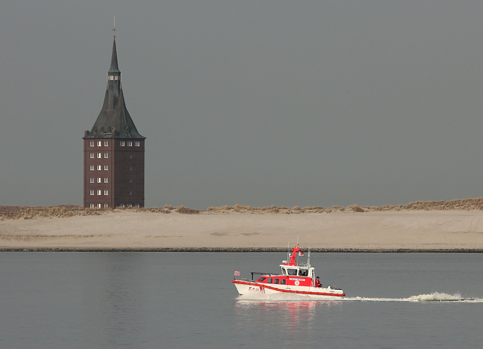 Seenotrettungsboot fährt vor Küste mit Leuchtturm im Hintergrund