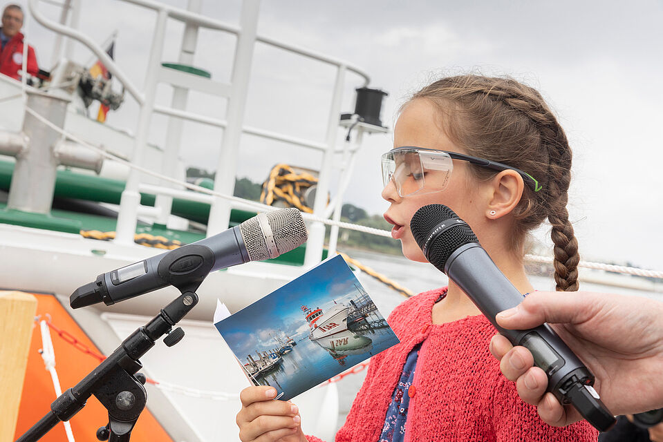 Kleines Mädchen mit Brille liest aus einer Karte und spricht in Mikrofone 