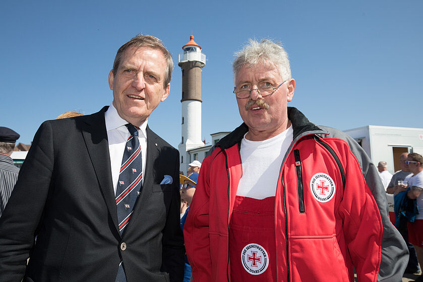 Ein Mann mit Anzug steht neben einem Ehrenamtlichen der DGzRS, im Hintergrund sieht man einen Leuchtturm