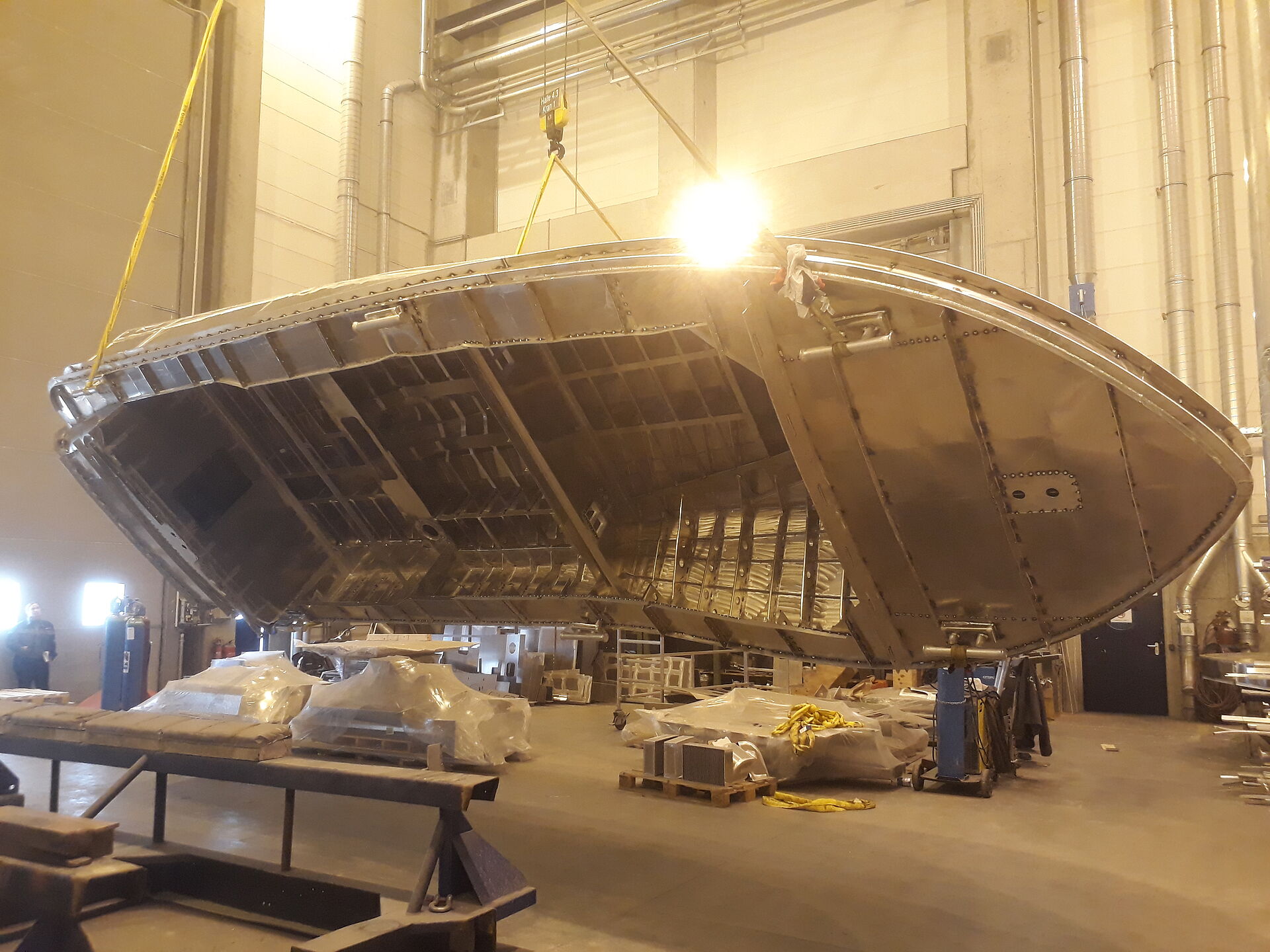Der Rohbau aus Aluminium eines Seenotrettungsbootes wird in einer Werfthalle von einem Hallenkran um die Längsachse gedreht.