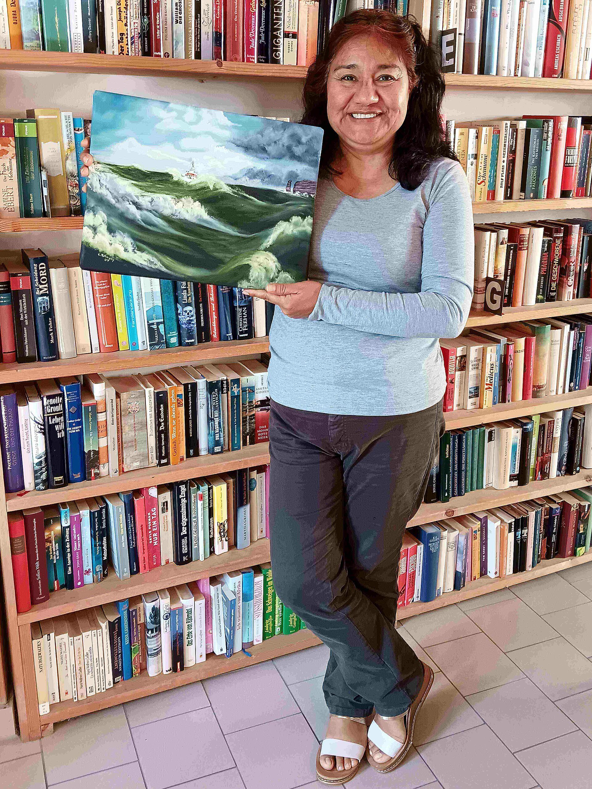 Eine Frau steht mit einem selbstgemalten Gemälde, Öl auf Leinwand, in der Hand vor einer Bücherwand. Das Bild zeigt einen Seenotrettungskreuzer bei schwerer See.