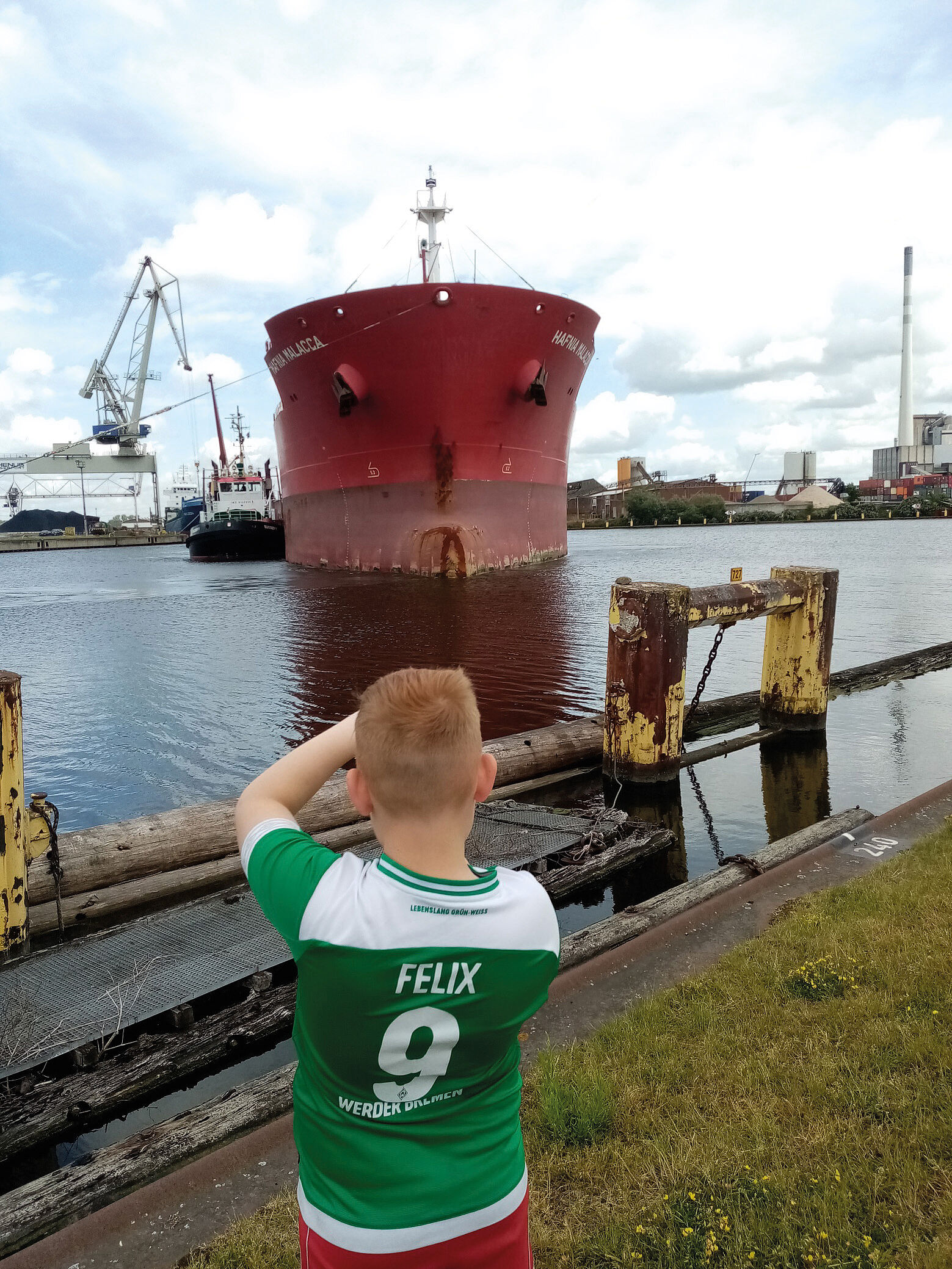 Kleiner Junge mit grünen Tshirt macht Fortos von einem großen Containerschiff, das frontal vor ihm im Wasser steht