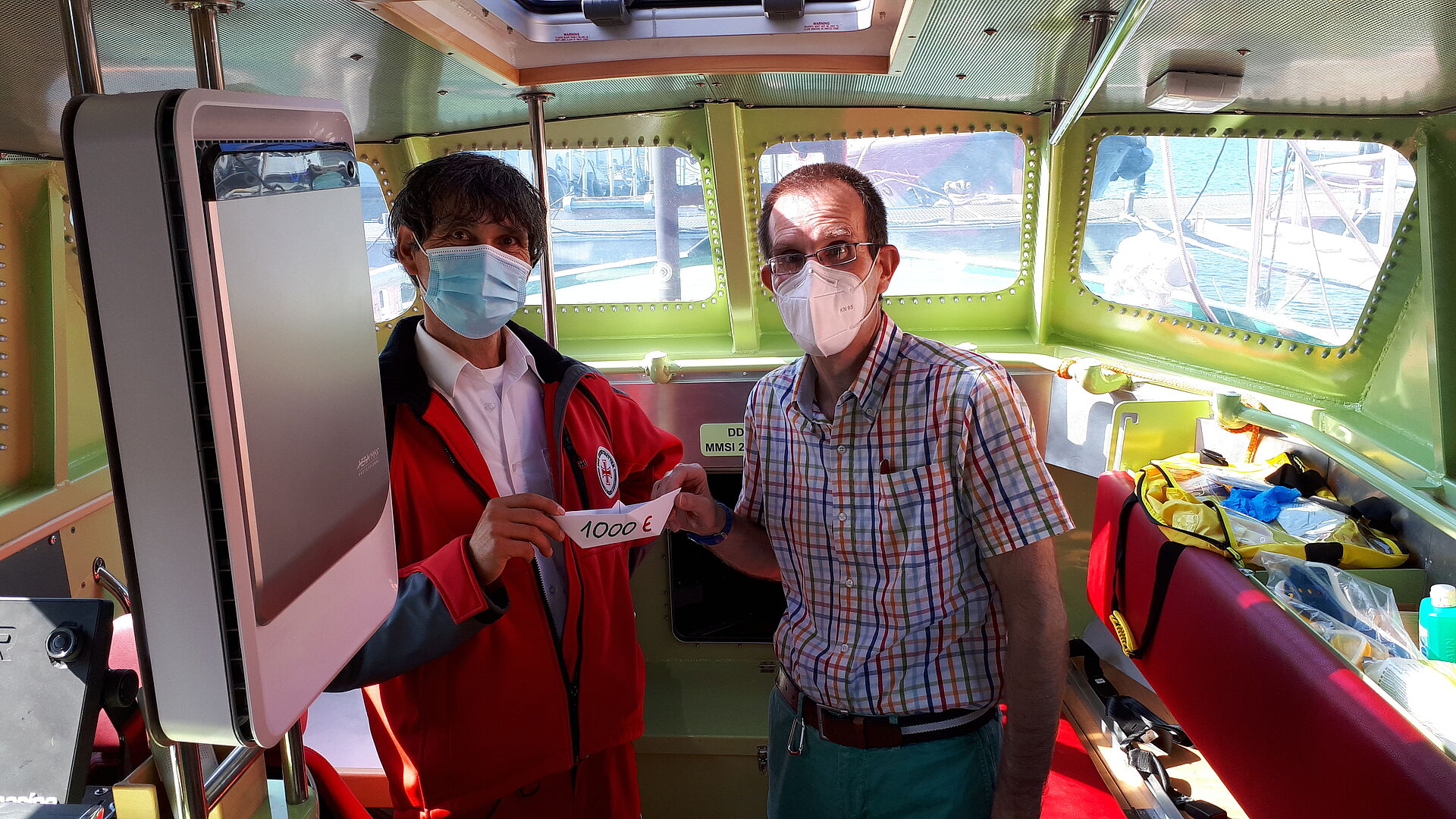 Zwei Männer mit Maske halten ein kleines Papierboot mit der Aufschrift "1000€"