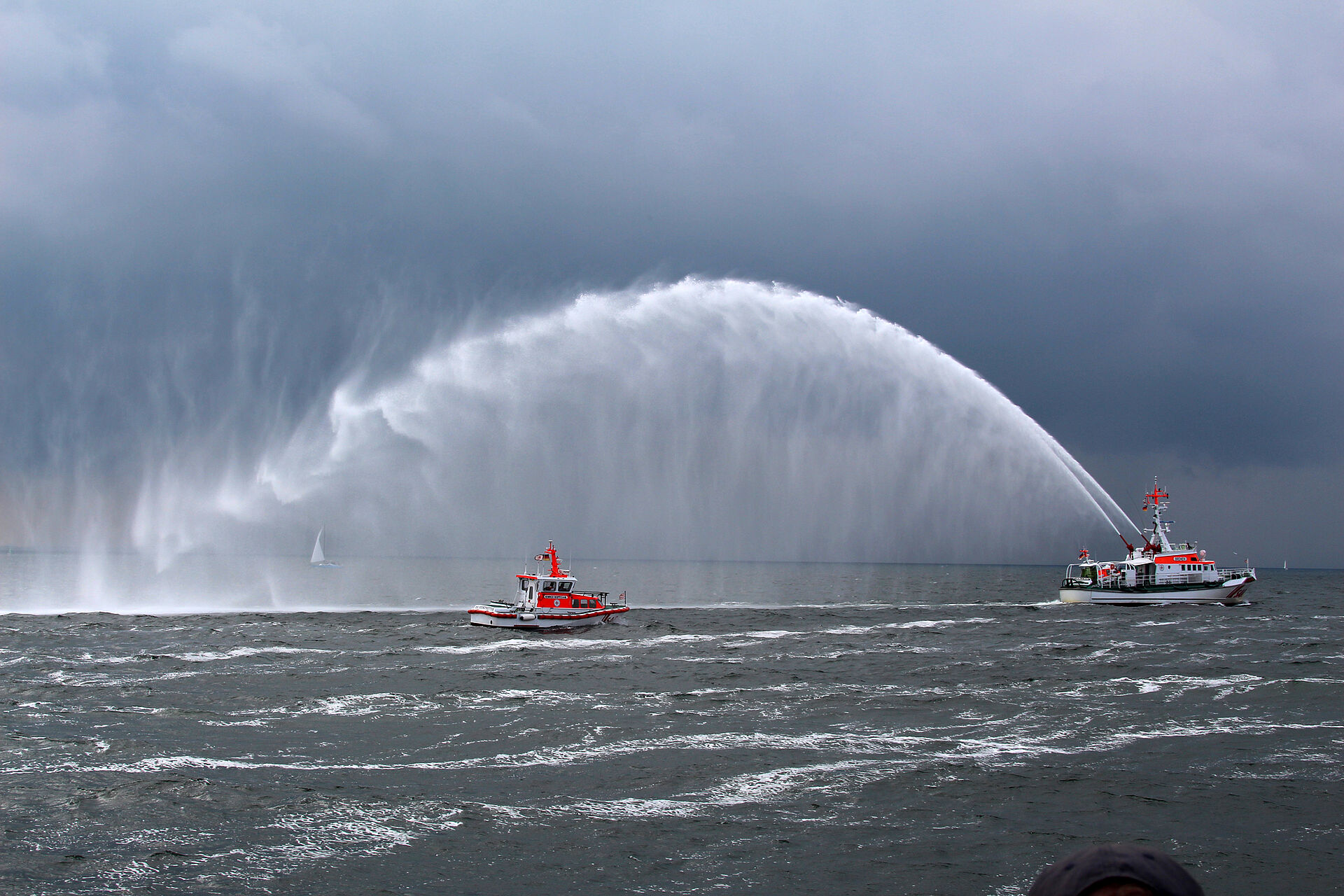 Ein Seenotrettungskreuzer wirft mit den Feuerlöschmonitoren hohe Wasserbögen, dahinter fährt ein Seenotrettungsboot
