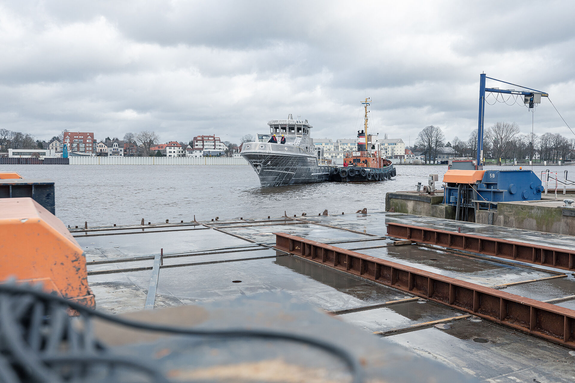 Dem Werftgelände nähert sich bei regnerischem Wetter der Aluminium-Rohbau eines Seenotrettungskreuzers, der auf der Weser schwimmt und von einem Hafenschlepper auf dem Wasser geschoben wird.