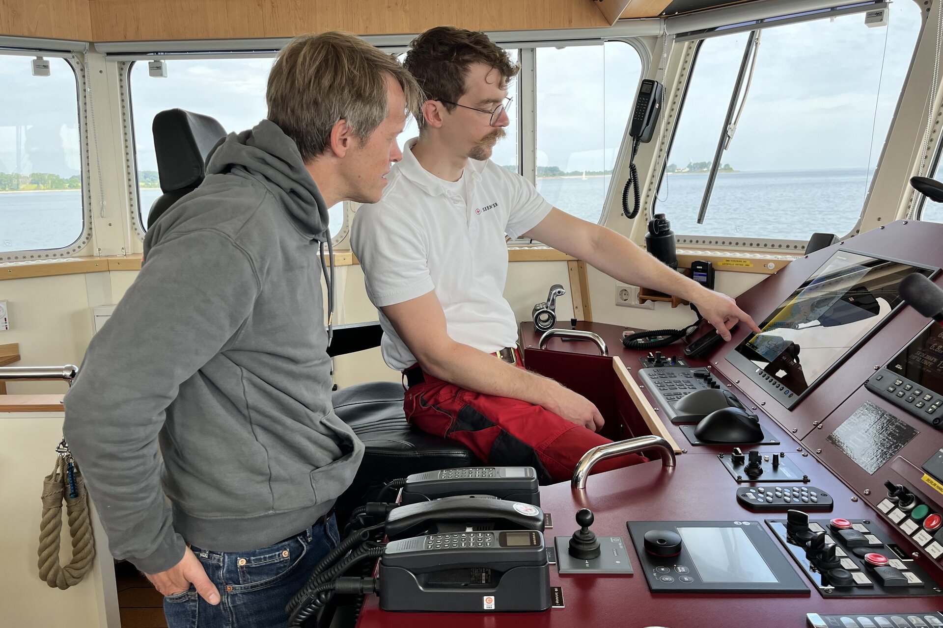 Zwei Personen stehen auf der Brücke eines Seenotrettungskreuzers vor den Navigationsgeräten. Eine Person sitzt und zeigt auf einen Bildschirm, während die andere neben ihr steht und aufmerksam auf die Anzeige blickt. Durch die Fenster ist ruhiges Wasser und eine Küstenlandschaft im Hintergrund zu sehen.. Zur Seite: Wetterprognosen helfen Seenotrettern auf See