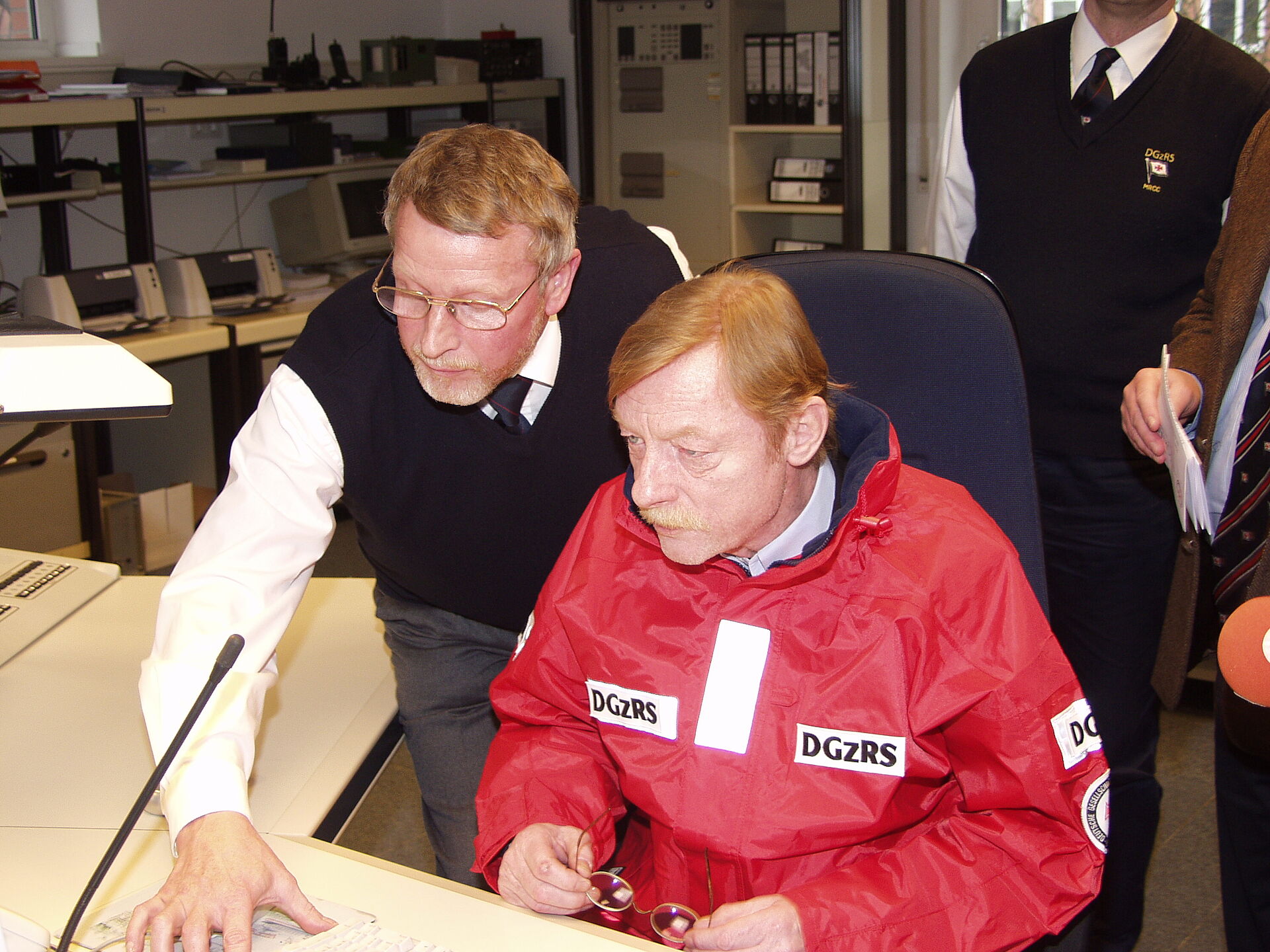 Schauspieler und DGzRS-Botschafter Otto Sander sitzt in roter Seenotretter-Jacke in der Seenotleitung Bremen. Ein Mitarbeiter der Seenotleitung zeigt ihm etwas an den Computern.