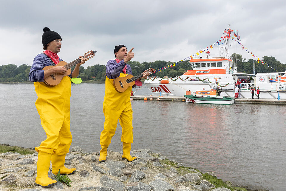 Zwei Seemänner mit gelber Gummihose und Gitarre machen Musik am Ufer während im Hintergrund ein Seenotretterboot schwimmt