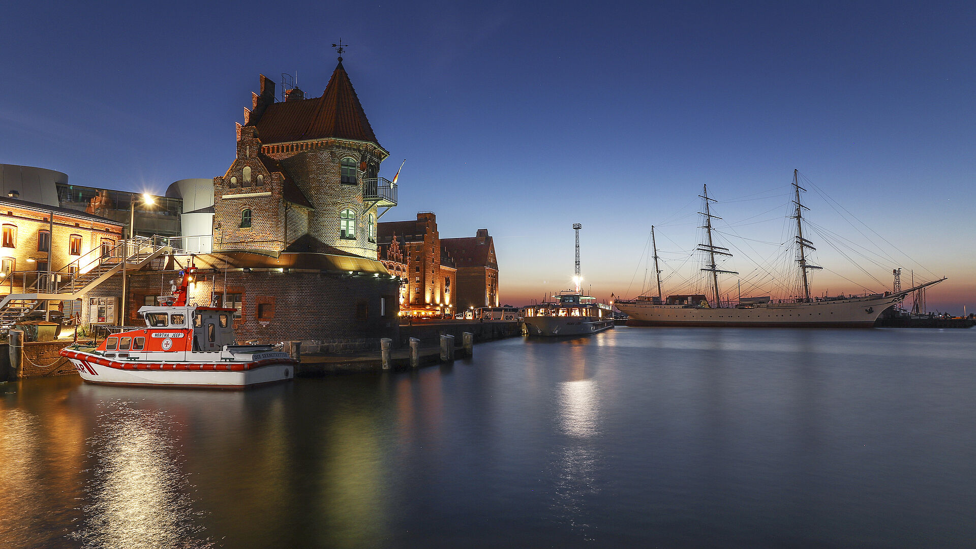 Sonnenuntergang im Hafen Stralsund mit historischer Kulisse. Das Seenotrettungsboot HERTHA JEEP liegt vor einem alten Lotsengebäude.