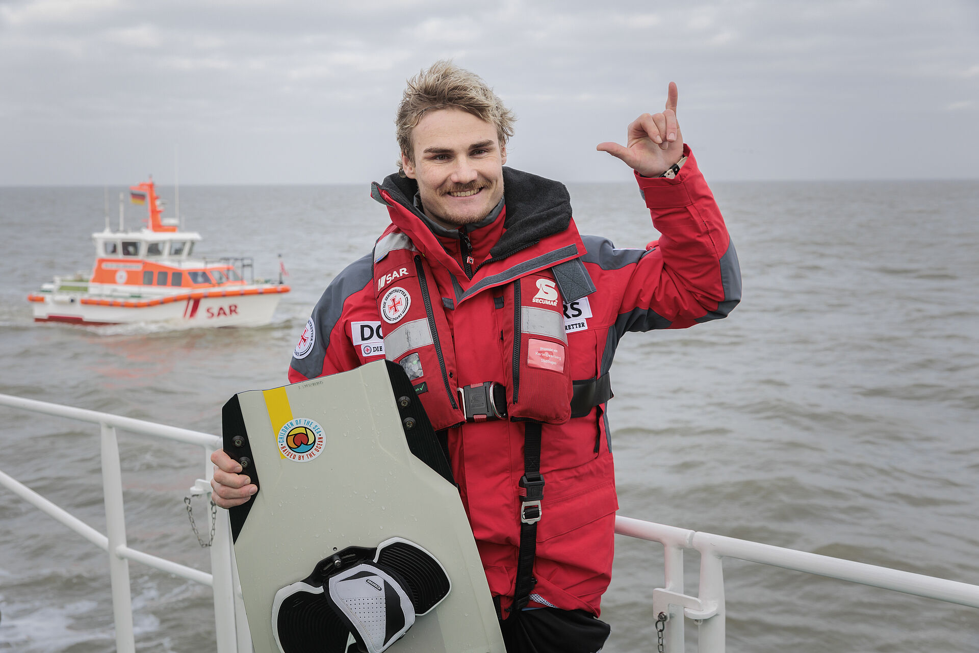 Kiteprofi steht mit seinem Board vor einer Reling eines Seenotrettungskreuzers, im Hintergrund fährt ein Seenotrettungsboot auf dem Wasser.