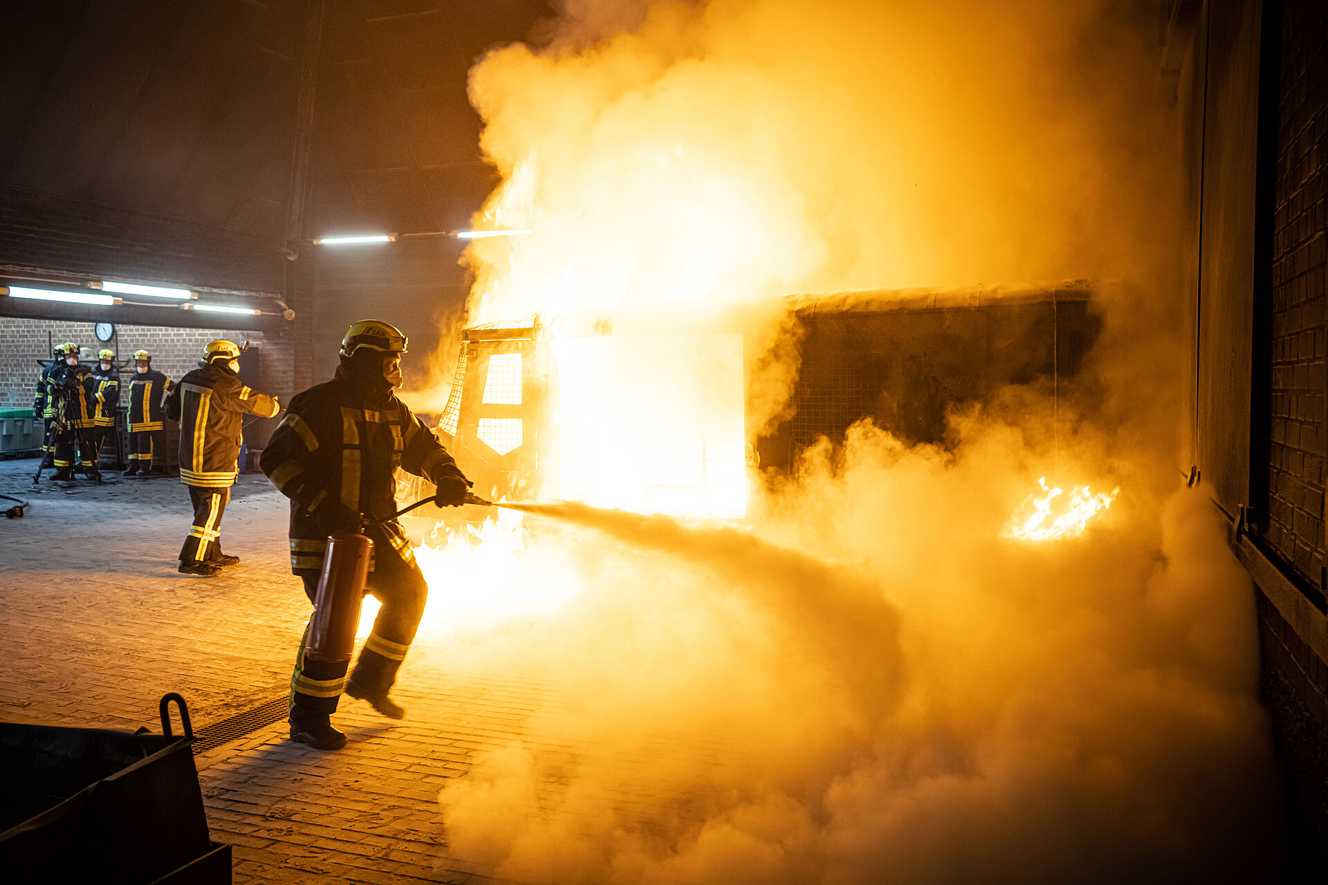 Zwei Personen mit schwerer Brandschutzkleidung löschen mit Handfeuerlöschern einen brennenden Kasko eines Hubschraubers in einer Halle.