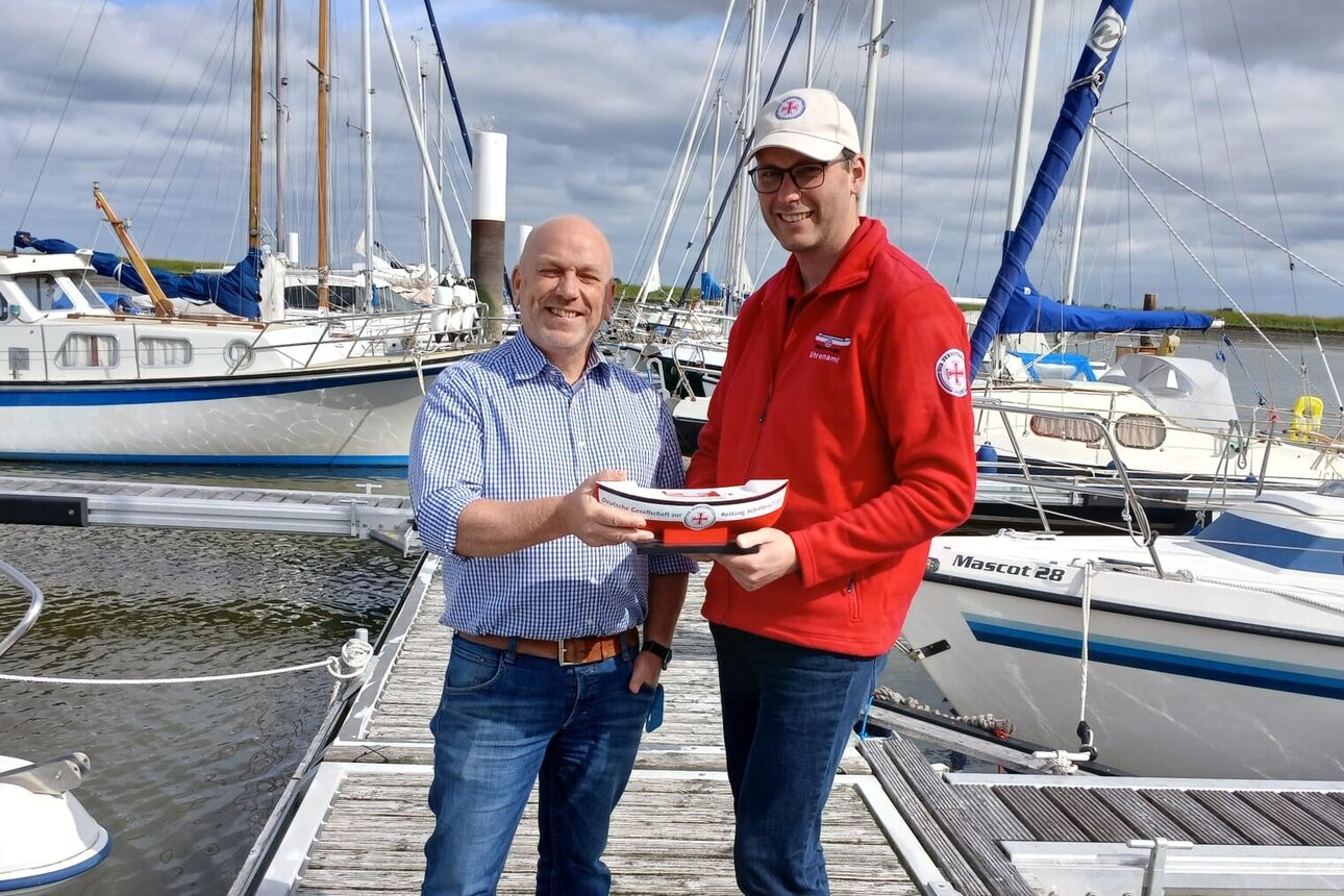 Zwei Personen stehen auf einem hölzernen Steg in einem Yachthafen zwischen mehreren Segelbooten und Motorbooten. Beide halten gemeinsam ein DGzRS-Sammelschiffchen vor sich in die Kamera. Eine Person trägt ein helles Hemd und Jeans, die andere die rote Dienstkleidung der Seenotretter und eine helle Kappe. Im Hintergrund sind dicht stehende Bootsmasten, vertäute Boote sowie ein bewölkter Himmel zu sehen.. Zur Seite: Spende im Andenken an einen Freund
