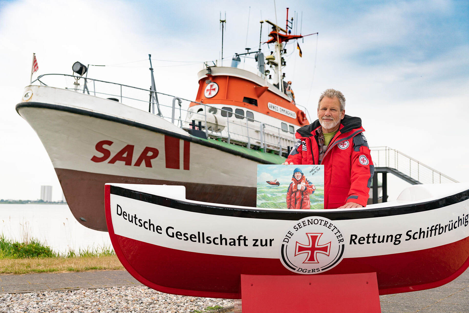 Cartoonist und Seenotretter-Botschafter Wolf-Rüdiger Marunde steht an einem großen Sammelschiffchen der DGzRS vor dem ehemaligen Seenotrettungskreuzer ARWED EMMINGHAUS auf Fehmarn.