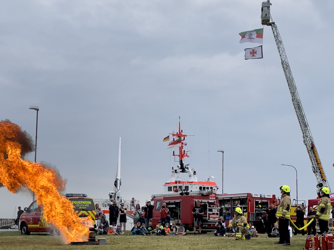Eine Flamme bei einer Übung, im Hintergrund eine Leiterwagen und andere Feuerwehrfahrzeuge