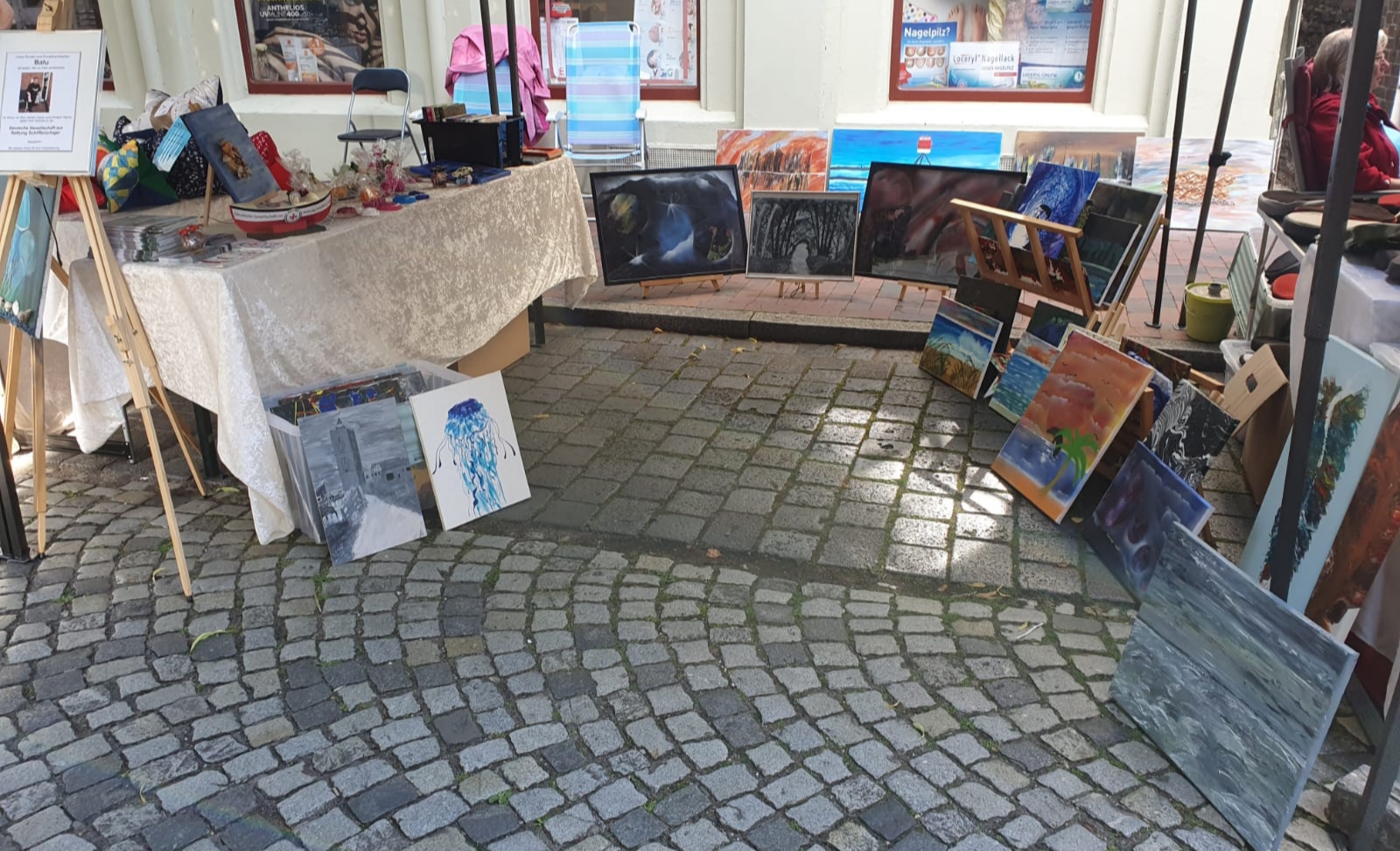 Auf dem Kunsthandwerkermarkt in Leer stehen maritime Bilder vom verstorbenen Reinhard Baalmann zum Verkauf, dessen Porträt auf einer Staffelei ausgestellt ist. Der Erlös der Verkäufe wird der DGzRS gespendet, das Sammelschiffchen steht auf einem der Tische, an dem die Werke lehnen.