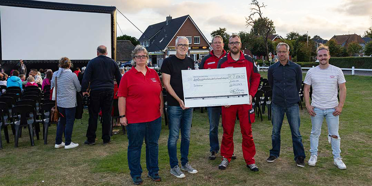 Auf einer Grünfläche stehen vor einer Kinoleinwand mit davorsitzendem Publikum die DGzRS-Ehrenamtliche Stefanie Jensen, Kinobetreiber Ralf Thomsen, die Seenotretter Lars-Peter Jensen und Thomas Berndt sowie Thomsens Kollegen Thorsten Ertel und Malvin Keppler im Abendlicht. Der Kinobetreiber sowie die beiden Seenotretter halten gemeinsam einen großen symbolischen Spendenscheck vor sich. 