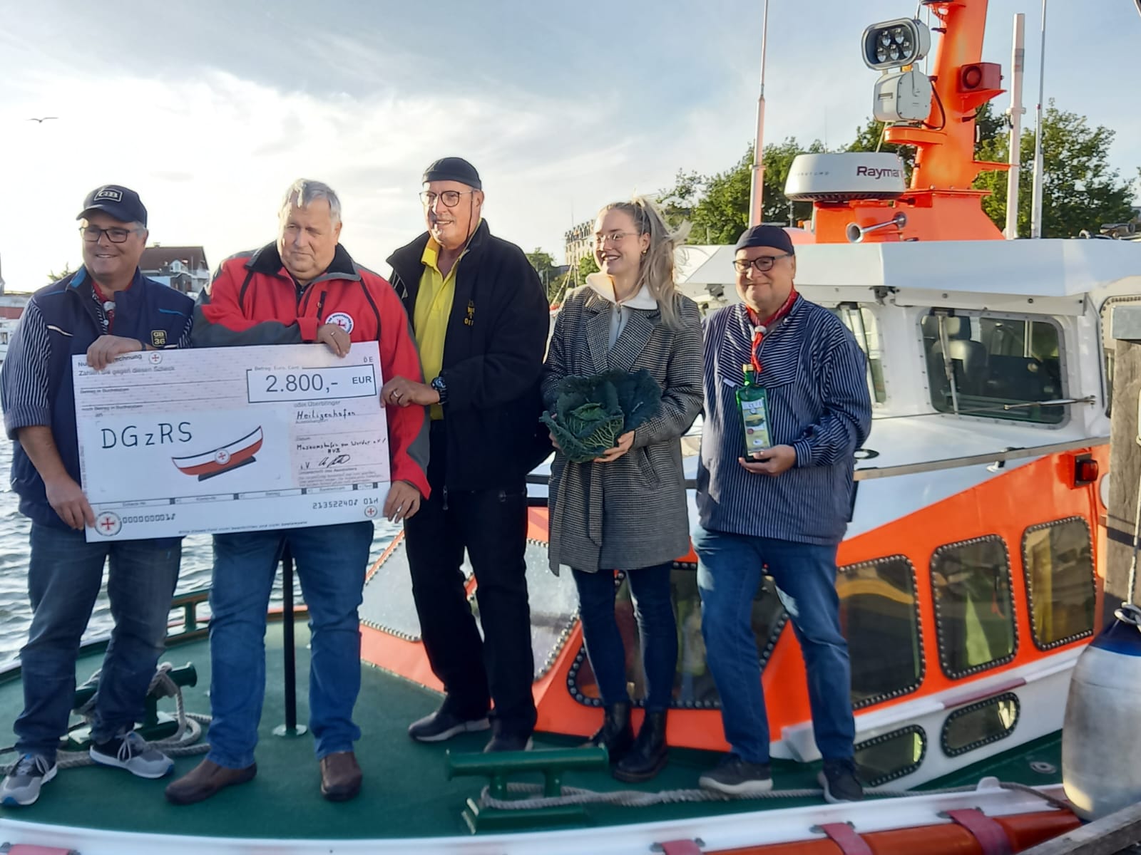 Auf dem Seenotrettungsboot HEILIGENHAFEN nimmt der freiwillige Vormann Frank Lietzow (2. v. l.) einen symbolischen Spendenscheck von dem links von ihm stehenden Herrn an. Rechts von ihm stehen drei weitere Organisatoren der „Maritimen Kohltage“ , darunter in der Mitte eine Frau, die einen Kohlkopf in den Händen hält.